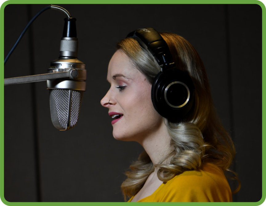 A narrator, a woman with shoulder length blonde hair, wears black headphones and a yellow top, as she narrates described video. The narrator stands in a recording booth and speaks into a microphone.